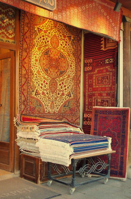 A display of various intricately patterned Persian and oriental rugs hanging vertically on a rack inside a shop. The rugs feature rich colors including red, green, gold, and blue, with detailed floral