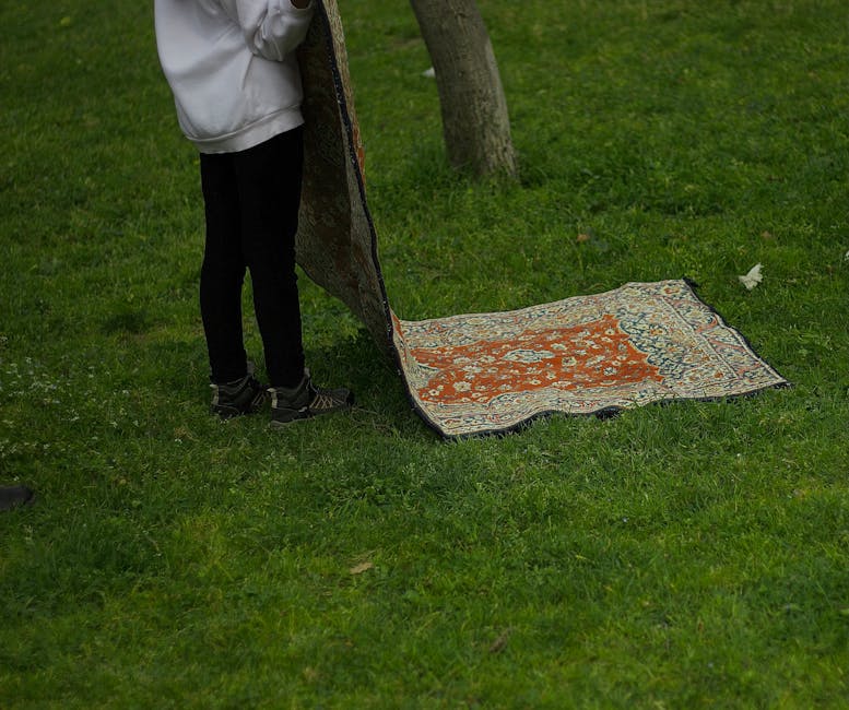 A person standing outdoors on a grassy area, holding a partially rolled-up decorative rug with an intricate floral pattern in shades of orange, beige, and blue. The rug is being aired or cleaned on a